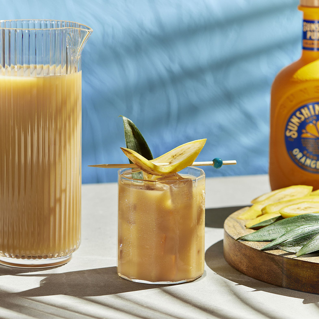 Cocktail with banana garnish next to a bottle of orange juice on a table with a blue background