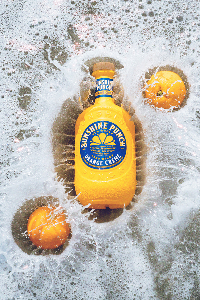 Sunshine Punch Orange Creme bottle with oranges splashing into the water on the beach
