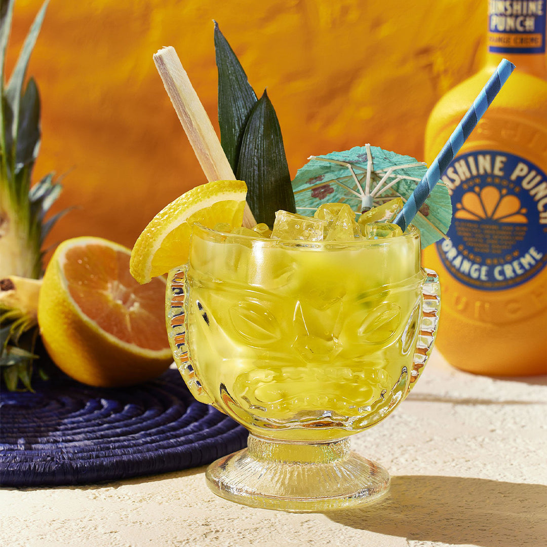 Cocktail in a glass with a straw, garnished with a lemon wedge and pineapple leaf, next to a bottle of Sunshine Punch.